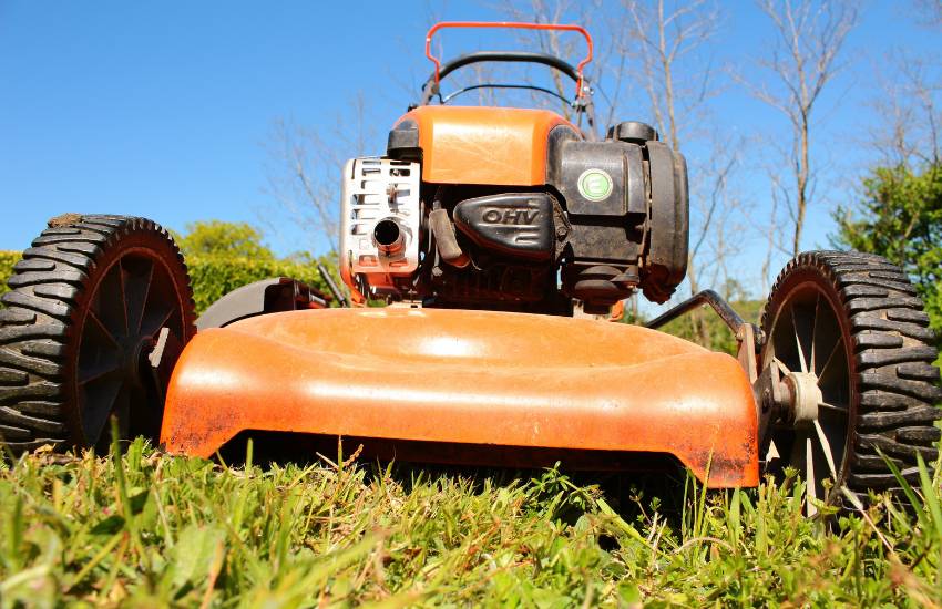 A low angle close-up of an orange gas-powered lawnmower sitting on green grass with a blue sky background.