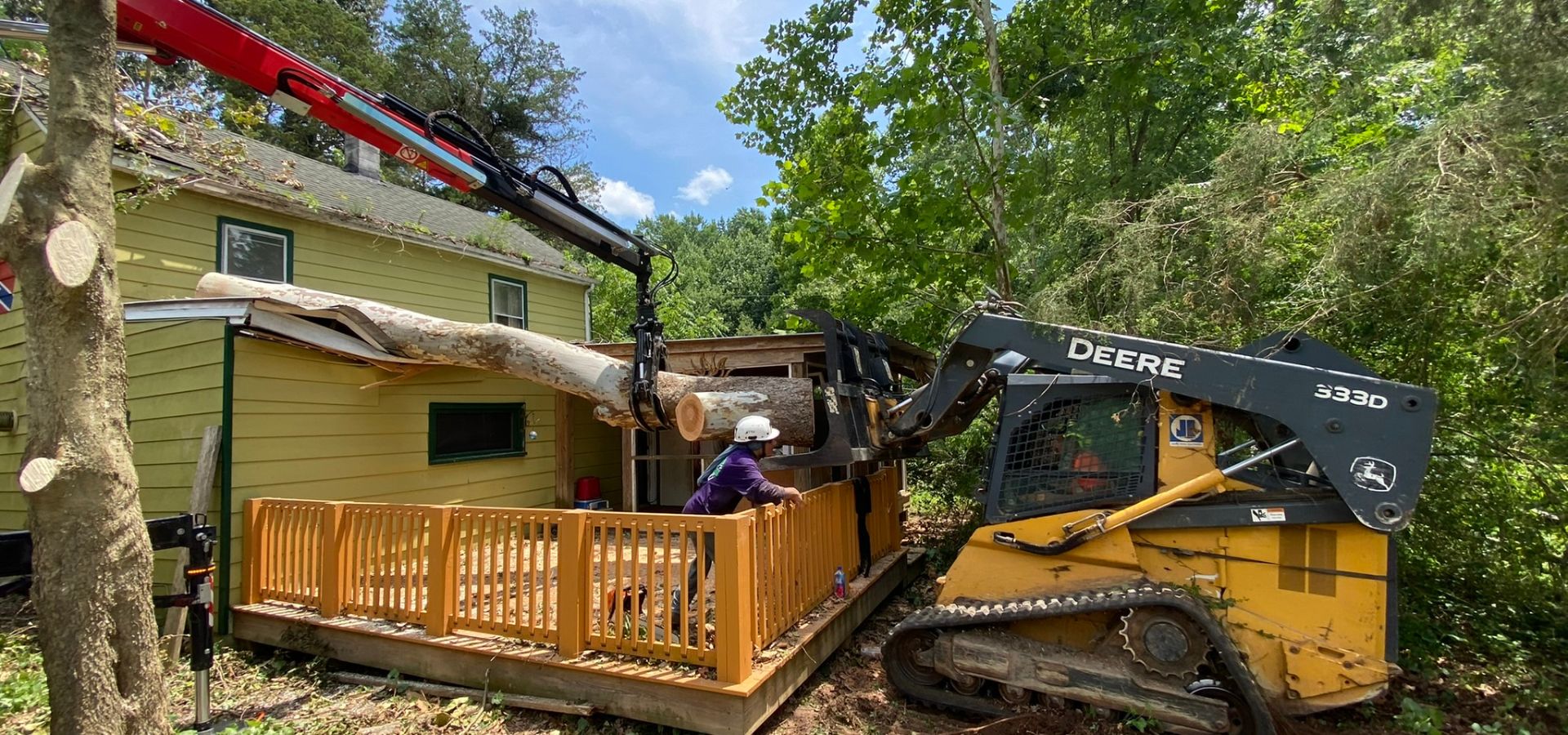 A yellow and black skid steer lifts a large section of fallen tree that’s fallen on a house and deck with some help from a grapple arm on a knuckle boom.